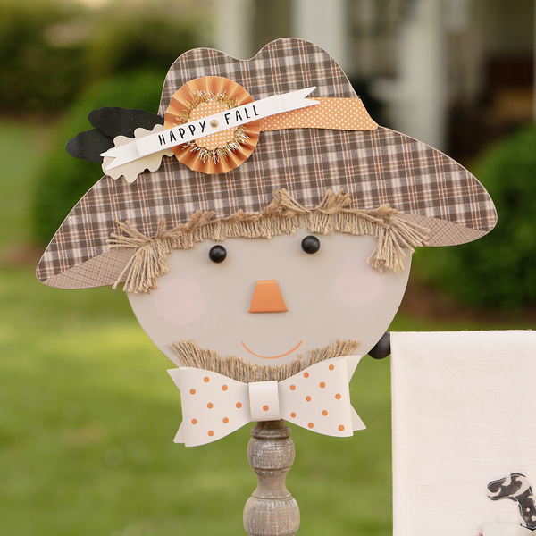 Decorative scarecrow with a plaid hat and 'Happy Fall' sign, standing outdoors.