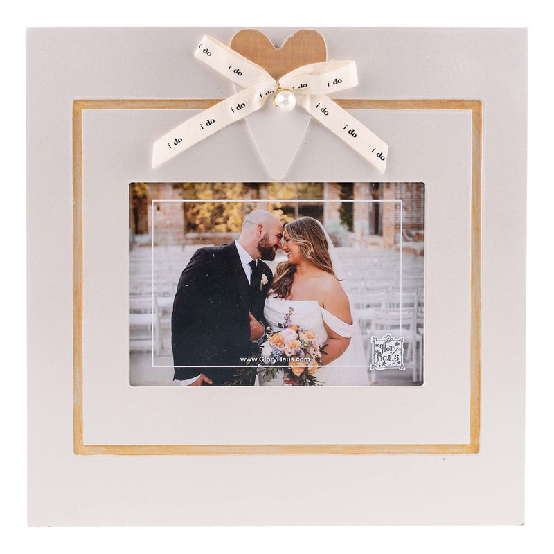 A square white photo frame with a light wood border, decorated with a white ribbon bow at the top. The ribbon is printed with the word 'i do' repeatedly and has a small pearl bead at the center.