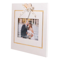 A square white picture frame with a gold inner border, decorated with a cream ribbon bow and heart topper. The frame is shown holding a wedding photograph of a couple, set against a plain white background.