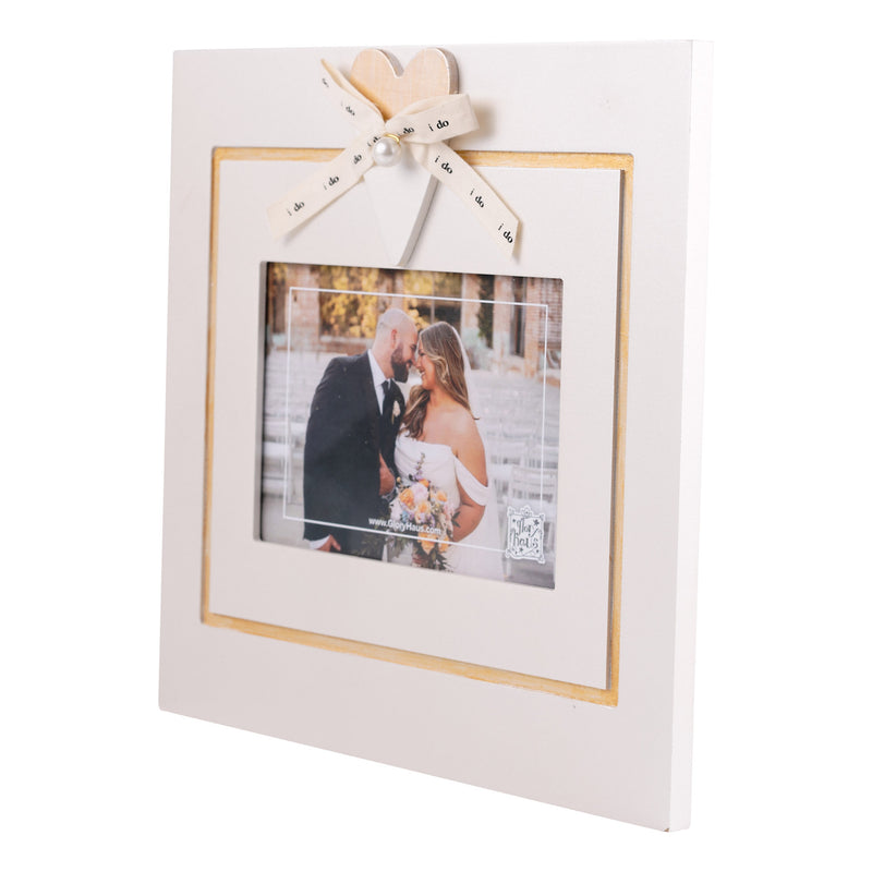A square white picture frame with a gold inner border, decorated with a cream ribbon bow and heart topper. The frame is shown holding a wedding photograph of a couple, set against a plain white background.