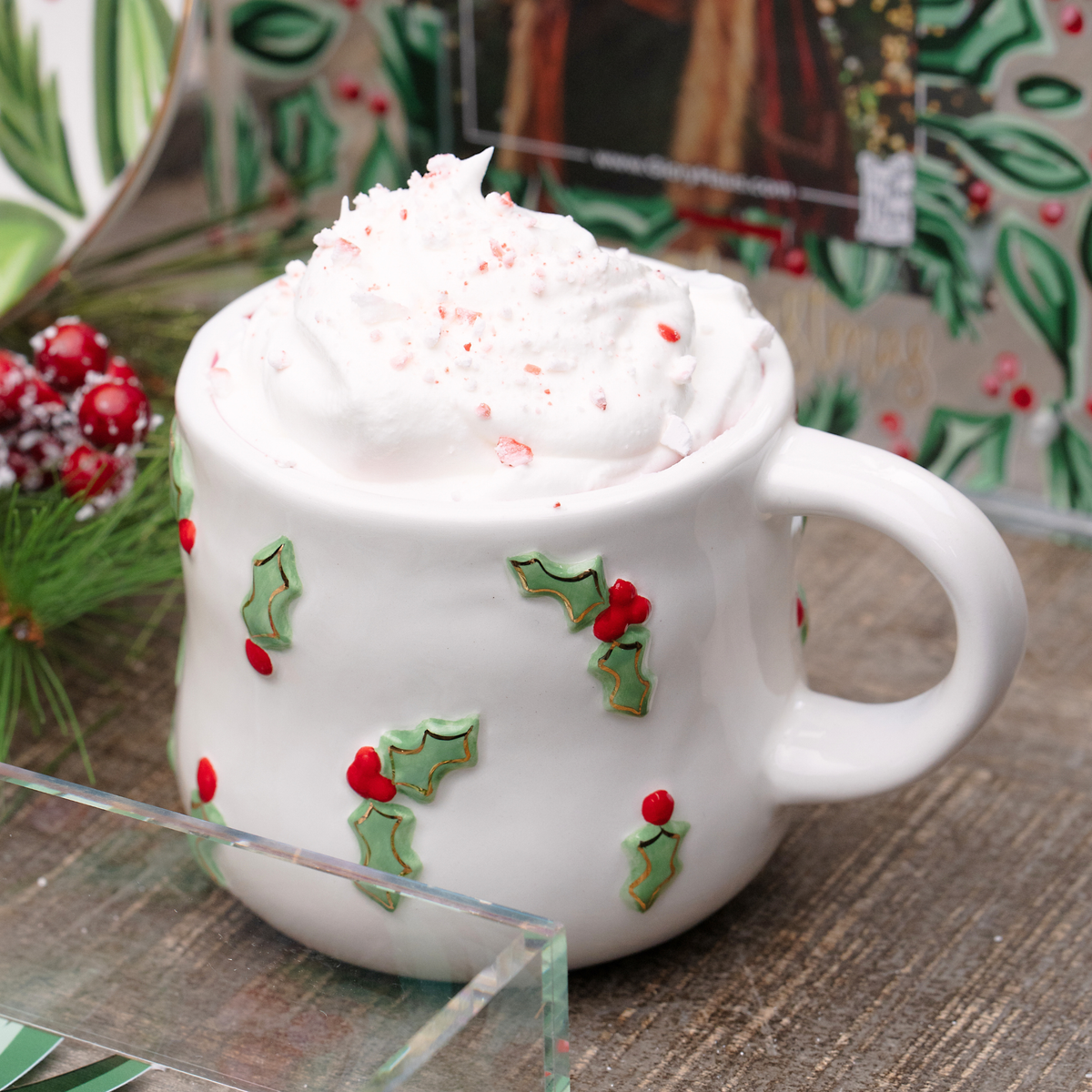 White mug with holly design filled with hot chocolate and whipped cream, set against a festive background.