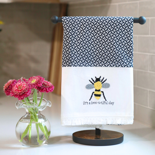 A black and white geometric patterned tea towel with a central bee design and the phrase 'it's a bee-u-tiful day' in yellow and green colors.